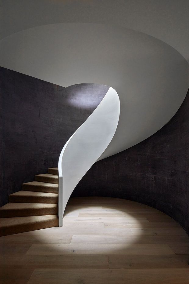 A sculptural curved staircase in a dark, rounded stairwell. A smooth white helical balustrade or stair wall wraps around the stair, contrasting with deep charcoal textured walls and pale timber flooring. The composition is minimalist and dramatic, with soft lighting emphasizing the sweeping curves and material contrast.