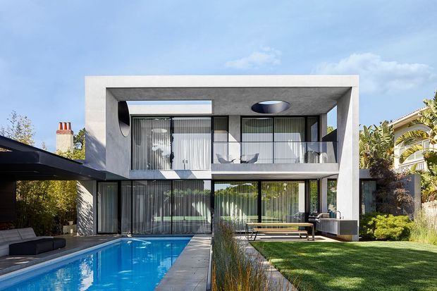 Exterior view of a contemporary two-storey residence with a strong rectangular concrete frame, extensive floor-to-ceiling glazing, upper-level balcony, and a swimming pool running alongside the house. The landscaped yard includes lawn, ornamental planting, and outdoor dining or lounge furniture. A covered carport is visible at left, with neighbouring residential context and tropical vegetation in the background.