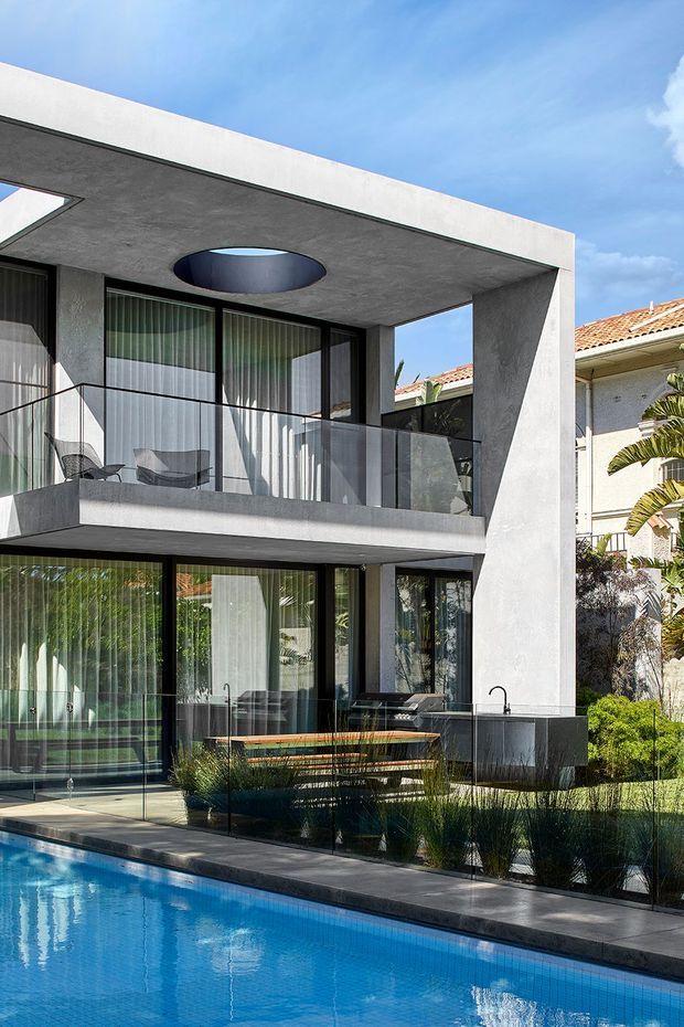 Modern two-storey residence viewed from the pool, featuring a projecting flat-roofed concrete terrace with a circular skylight, dark-framed floor-to-ceiling glazing, a glass balustrade, and a compact outdoor dining area beside the pool. The facade is rendered in light grey and white, with landscaped planting and a neighbouring tiled-roof house visible in the background.