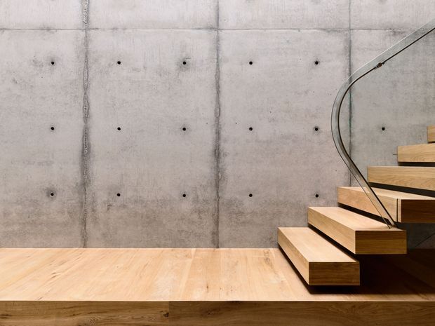 Minimalist interior detail featuring a floating timber staircase with a curved clear glass balustrade against a smooth, board-formed concrete wall. The stair and adjacent floor platform use warm natural timber, while evenly spaced concrete form-tie marks and vertical seams add an industrial architectural character.