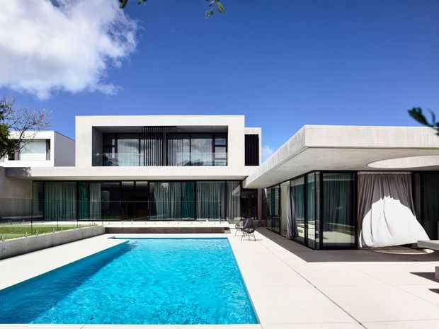 A bright modern flat-roof residence arranged around a rectangular swimming pool. The home features crisp white rendered volumes, extensive floor-to-ceiling glazing, dark window frames, covered outdoor areas, and a paved pool terrace under a clear blue sky.