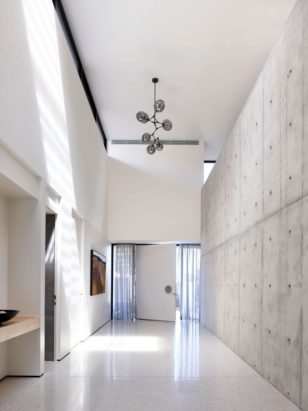 A bright, minimalist double-height interior hallway or entry space with polished light terrazzo flooring, a raw exposed concrete feature wall, white walls and built-in joinery. Floor-to-ceiling glazing with sheer curtains brings in strong daylight, while a sculptural dark metal pendant hangs from the high ceiling. A large pivot door and artwork are visible at the far end.