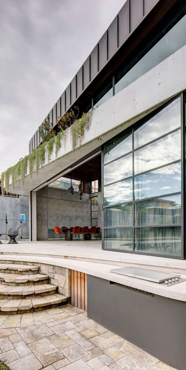 A modern multi-level concrete building exterior with a sheltered open-air dining terrace, large industrial-style glazed doors and windows, planted greenery cascading from an upper planter, and broad stone-paved steps and courtyard paving. Exposed concrete, dark metal framing, timber soffits, and red dining chairs create a contemporary urban architectural composition.