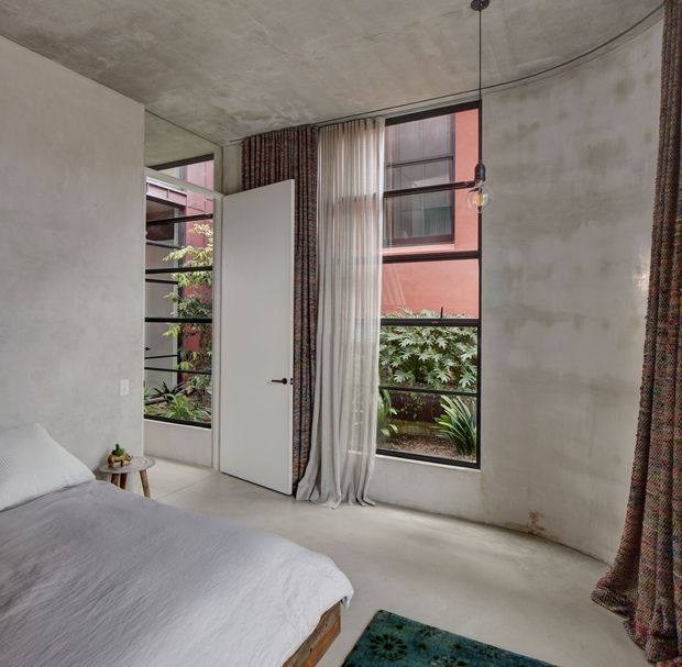 A minimalist bedroom with a low platform bed, pale bedding, raw concrete-look walls and ceiling, polished concrete flooring, and full-height steel-framed windows overlooking a planted courtyard. Sheer and patterned floor-length curtains soften the space, while a simple exposed pendant light and small timber bedside stool add understated detail. A white door opens beside the bed.