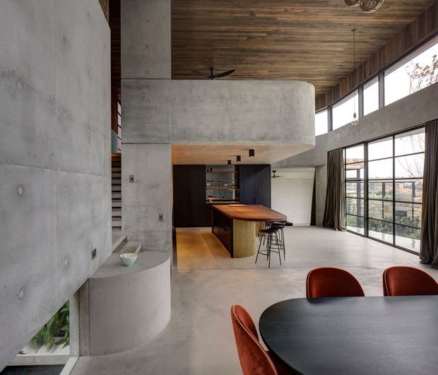 Open-plan contemporary interior combining a kitchen, dining area and living space. The room features exposed concrete walls and a sculptural curved concrete bulkhead, a timber-clad ceiling, polished concrete flooring, dark fixed kitchen joinery, a timber island with stools, and a large black dining table with rust-coloured upholstered chairs. Floor-to-ceiling glazing provides expansive urban views and abundant natural light.