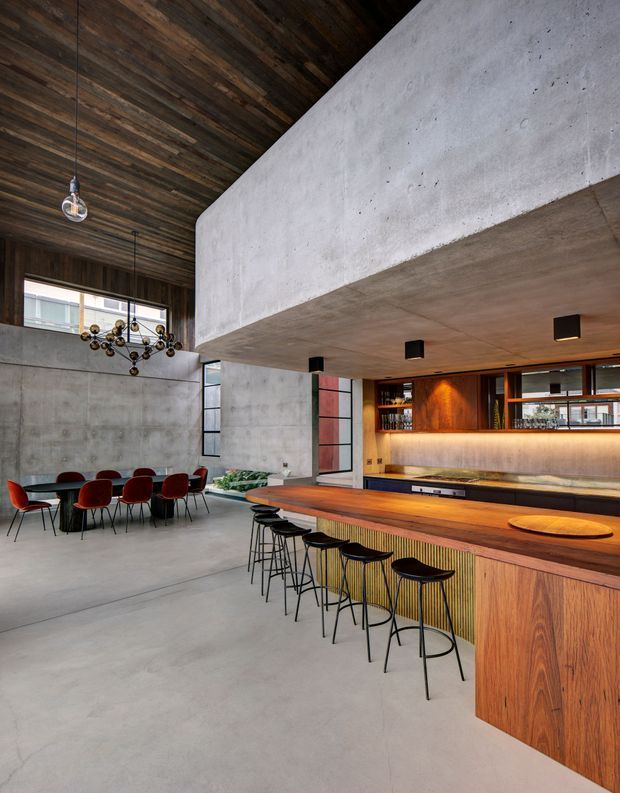 A dramatic open-plan interior combining a dining area and substantial home bar. The space features polished concrete floors and walls, a dark reclaimed timber ceiling, a large exposed concrete beam and soffit, warm timber bar cabinetry, a long stone-look benchtop, integrated shelving with bottles, and a row of black bar stools. A dining table with red upholstered chairs sits beneath a sculptural pendant light. The high ceiling, exposed structure, and restrained industrial palette create a refined, atmospheric hospitality-style setting.
