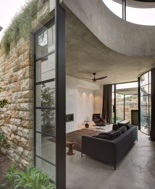 A sophisticated living room viewed through a tall black-framed glazed opening, with a dark sectional sofa, leather lounge chair, rug, and small fireplace. The space features a curved exposed concrete ceiling and balcony-like form, polished concrete flooring, white plaster walls, extensive glazing, and substantial rough stone masonry with planted greenery along the exterior edge.