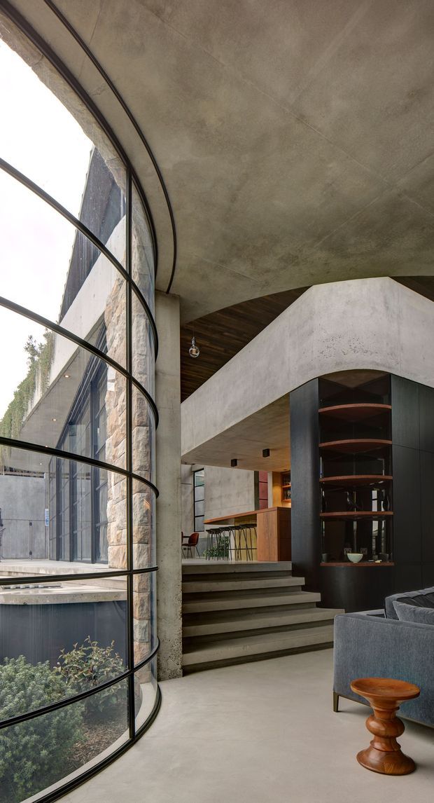 A dramatic contemporary interior with a tall curved glazed wall, exposed concrete ceilings and floors, stone-clad structural columns, and a sculptural staircase leading to a bar or kitchen area. Dark timber ceiling lining and black built-in shelving contrast with the concrete, while a grey sofa and timber side table occupy the foreground. The space has a strong indoor-outdoor connection and generous vertical volume.