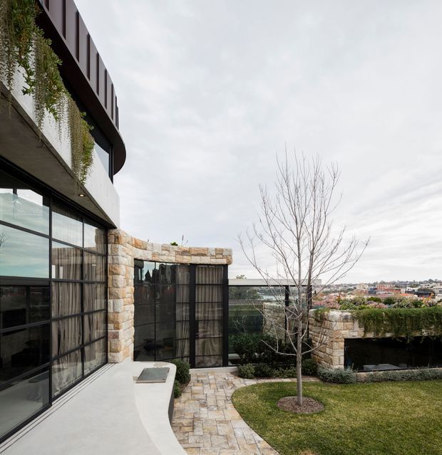Exterior view of a contemporary multi-level residence with extensive black-framed glazing, curved upper facade, pale stone masonry, planted facade elements, and a landscaped courtyard. A paved path curves through the lawn toward a glazed garden pavilion or enclosed outdoor area, with suburban buildings visible beyond.