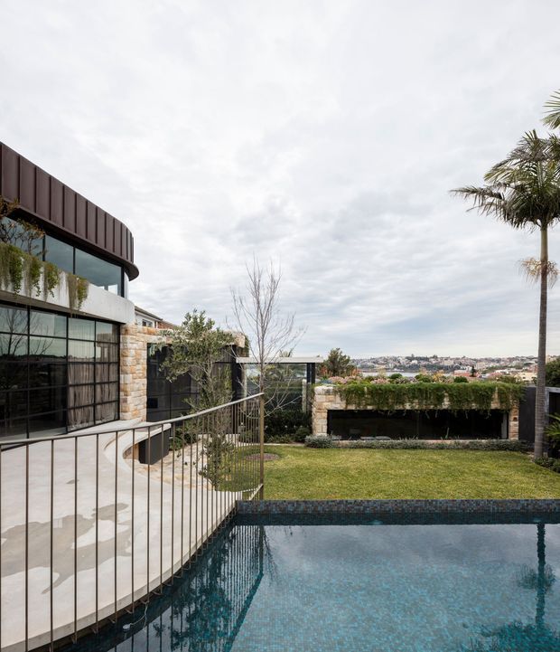 A contemporary multi-level residence viewed from an elevated outdoor terrace, featuring a swimming pool in the foreground, curved pale paving and metal balustrades, extensive dark-framed glazing, stone walls, a planted rooftop-like garden structure, and a broad suburban city outlook. A tall palm and landscaped lawn soften the modern architectural composition beneath an overcast sky.