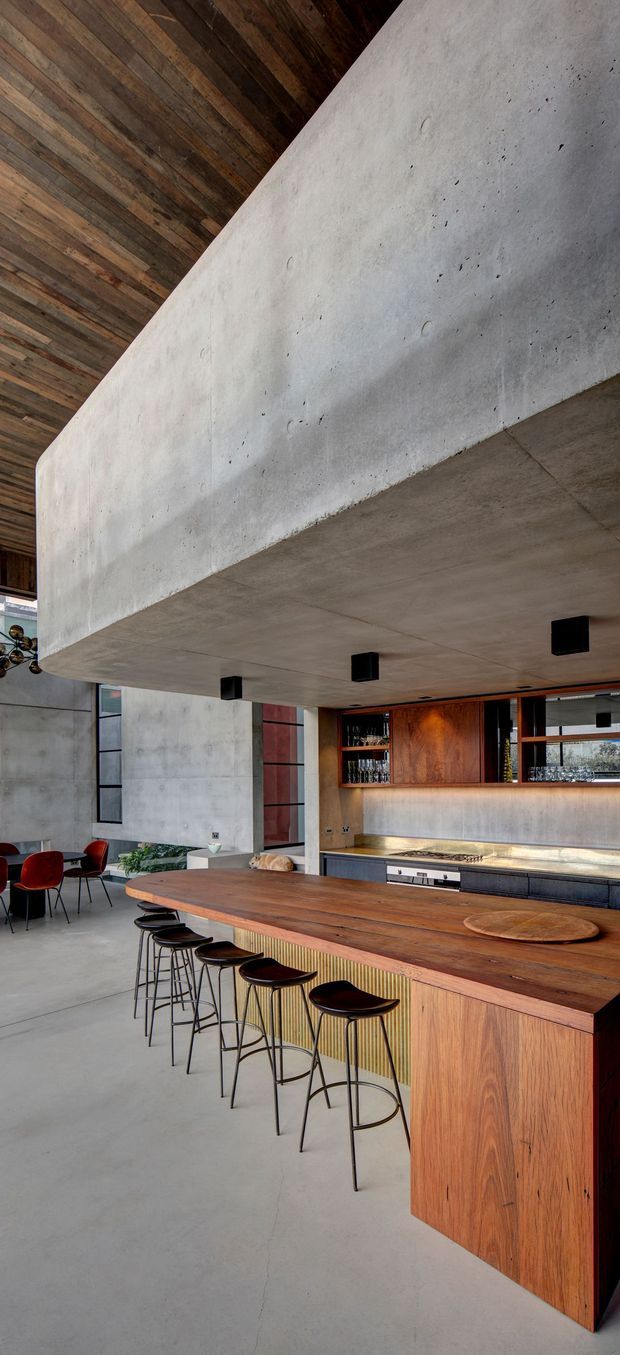 A dramatic contemporary open-plan kitchen and home bar with a long timber island, integrated seating, concrete flooring and walls, and a large projecting concrete ceiling volume. The bar features dark timber cabinetry, open bottle shelving, warm under-cabinet lighting, and a stainless-steel cooking area. A dining area with red upholstered chairs is visible in the background. The space has an industrial, refined character with strong exposed materiality and a high ceiling.
