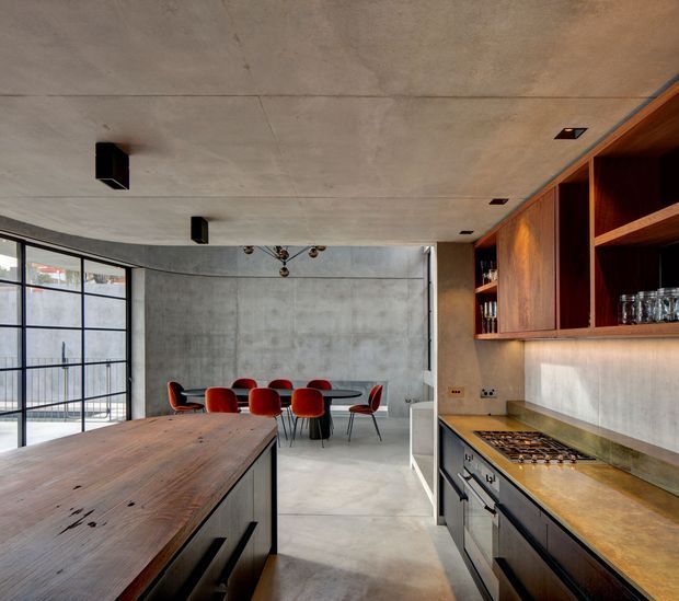 Open-plan industrial-style kitchen and dining area with a large timber island in the foreground, stainless-steel range and dark lower cabinetry along the right wall, warm timber open shelving and cupboards, exposed concrete walls and ceiling, and black-framed floor-to-ceiling glazing. A dining table with red upholstered chairs sits beyond the kitchen.