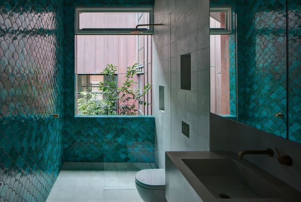 A compact contemporary bathroom with a wall-hung toilet and a dark integrated vanity basin. The walls are clad in a striking teal scallop-pattern mosaic tile, contrasted with large pale grey wall and floor tiles. A large mirror, narrow high windows, and indoor greenery bring light and depth to the space. The palette is dark teal, grey, and black with a bold patterned feature finish.