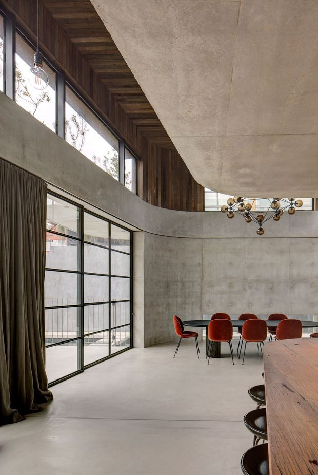 A dramatic double-height dining space with exposed concrete walls and floor, tall black-framed glazing, dark timber-lined ceiling and upper wall surfaces, and a curved concrete mezzanine or balcony edge. A long timber dining table is paired with rust-red upholstered chairs, while a sculptural brass-and-glass chandelier hangs centrally. Heavy dark curtains frame the glazing, creating an industrial yet refined interior.