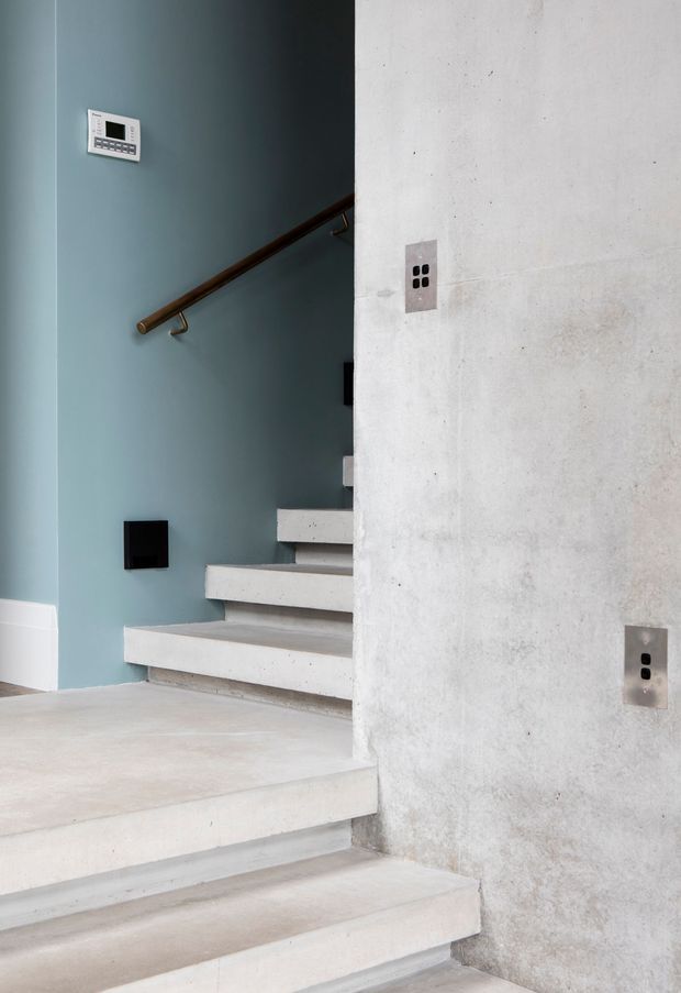 Interior view of a minimalist stairway with pale concrete steps and a rough light-grey concrete wall. The stairwell has a muted blue painted wall, a dark timber handrail with metal brackets, simple black electrical outlets and switches, and a small wall-mounted control panel. The composition highlights the modern material palette and clean geometric stair design.