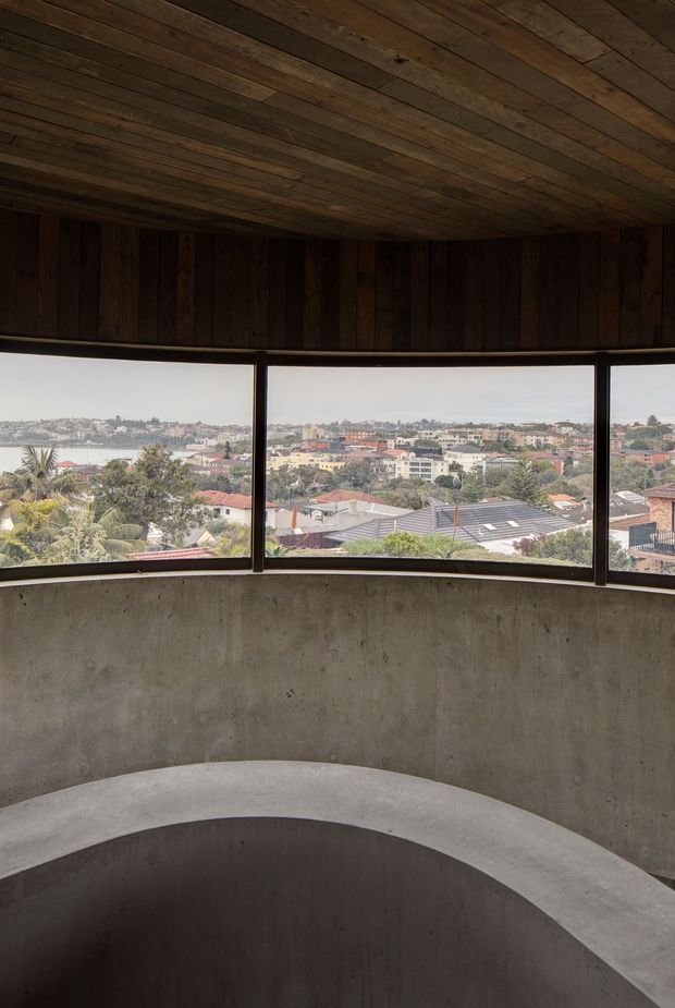 A curved interior landing or stairwell with a broad concrete balustrade and raw concrete walls. Dark timber boards line the ceiling and upper wall, while a continuous band of dark-framed windows provides expansive views over a dense coastal or waterside residential neighbourhood. The space has a raw, sculptural, materially expressive character.