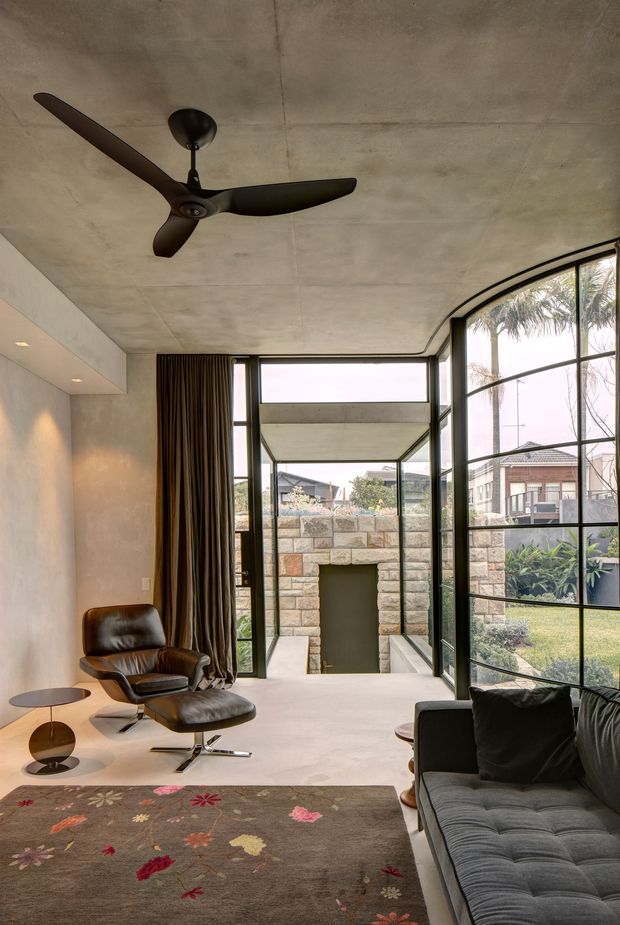 A refined modern living room with a raw concrete ceiling and smooth pale walls, furnished with a dark leather lounge chair and ottoman, a black sofa, low round side tables, and a floral patterned rug. A black ceiling fan hangs overhead. Full-height black-framed glazing and a curved glazed wall open toward a landscaped courtyard, with a stone-clad outdoor fireplace visible beyond.