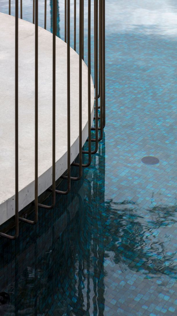 Close-up architectural detail of a swimming pool edge, showing blue mosaic pool tiles, a pale concrete slab or coping, and a dark metal vertical safety balustrade with curved supports descending into the water. The image is tightly framed around the pool structure and contains no visible room or wider property context.