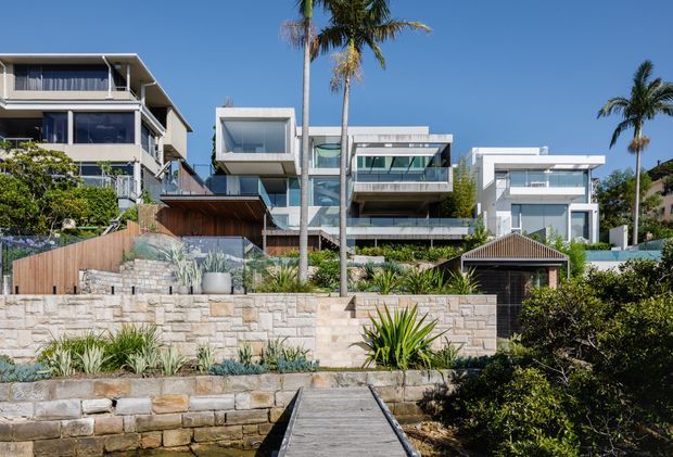 Exterior view of a contemporary multi-level waterfront residence, photographed from a timber jetty or boardwalk. The property features layered box-like volumes, extensive glazing, balconies, flat rooflines, pale stone retaining walls, timber fencing and decking, landscaped planting, and tall palms under a clear blue sky.