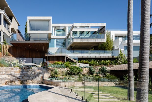 Exterior view of a multi-level contemporary residence on a landscaped, sloping site. The house features crisp white rendered volumes, extensive floor-to-ceiling glazing, projecting balconies, timber soffits and screens, and layered stone retaining walls. A swimming pool, lawn, low-water planting, glass pool fencing and tall palm trees occupy the foreground.