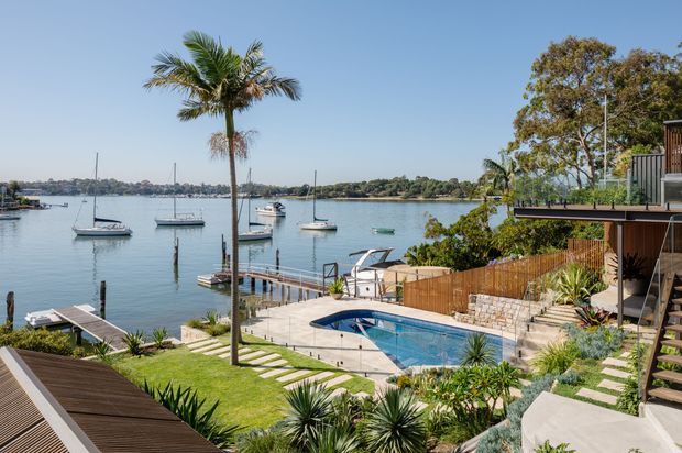 A landscaped waterfront residential property viewed from an elevated outdoor terrace or garden. The scene features a swimming pool, timber decking, stone retaining walls, stepped concrete paths, tropical and drought-tolerant planting, a timber privacy fence, a private dock, and a calm bay with moored sailboats. Part of a contemporary multi-level home with timber-clad outdoor structures is visible at right.