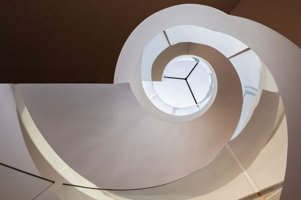 Upward-looking view of a sculptural spiral staircase or stairwell wrapping around a circular central opening with a round skylight or glazed oculus above. Smooth pale plaster surfaces contrast with dark brown ceiling areas, and a simple black clock-like radial element is visible at the centre. The image is dominated by the stairwell geometry and curved architectural form.