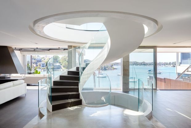 Bright contemporary interior dominated by a sculptural curved spiral staircase with dark timber treads, a translucent glass balustrade, and a sweeping white underside rising through a large circular ceiling opening. Full-height glazing provides expansive waterfront views, while polished pale flooring, light walls, and minimal furnishings create an airy, refined setting.