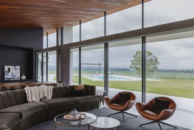 A spacious contemporary living room with a large dark sectional sofa, two brown leather lounge chairs, round glass coffee tables, and a dark textured rug. Full-height glazing opens to a broad landscaped lawn and distant rural landscape, while a warm timber-lined ceiling and dark built-in wall create contrast. The room has a refined, relaxed atmosphere and strong indoor-outdoor connection.
