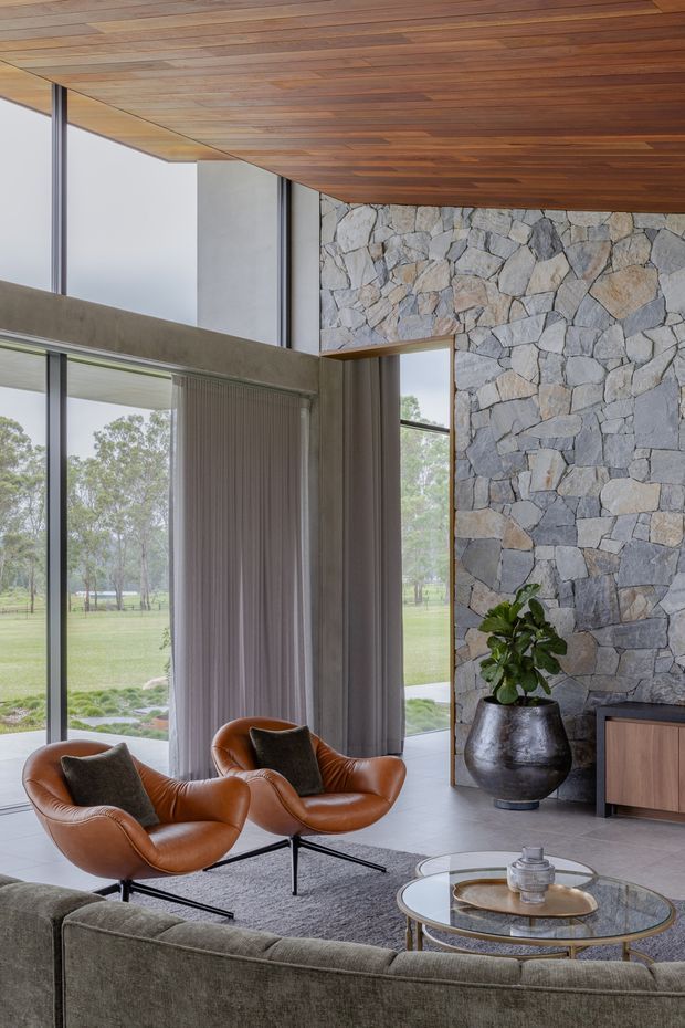 A refined living room with full-height glazing overlooking a green landscape. The space features a dramatic rough stone feature wall, warm timber-lined ceiling, concrete structural elements, grey curtains, a muted grey sofa, cognac leather lounge chairs, and layered round glass coffee tables. The room has a high ceiling and a strong indoor-outdoor connection.