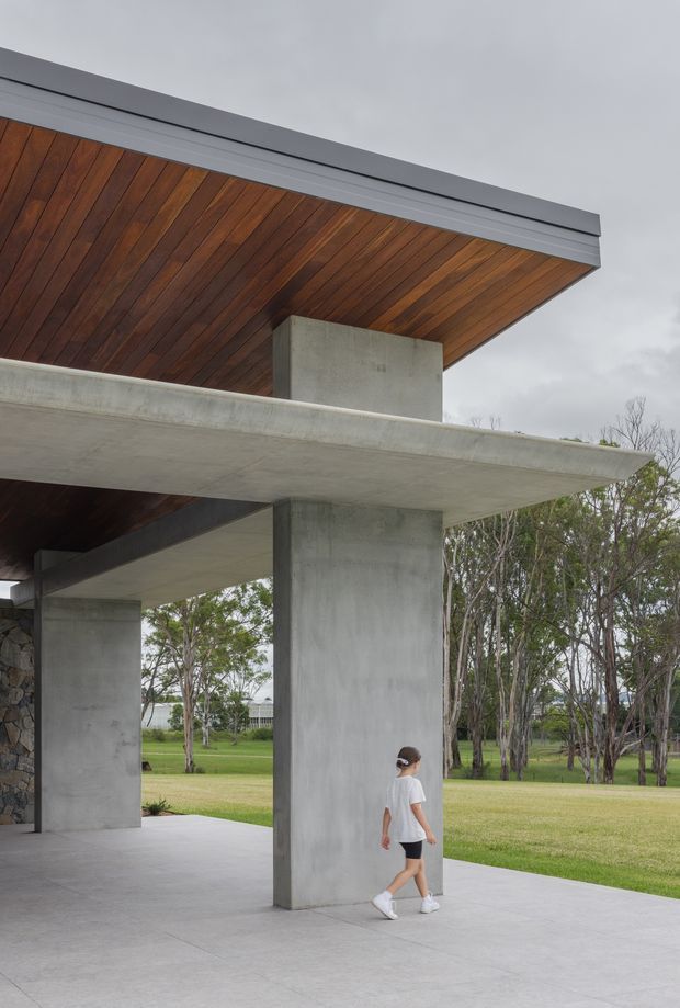Exterior architectural photograph of a modern pavilion-like building with broad projecting concrete slabs, tall raw concrete columns, a timber-lined soffit, dark metal roof edging, and partial stone wall construction. The open ground-level structure frames a landscaped lawn and trees beyond, with a person providing scale. Overcast natural light emphasizes the restrained material palette and strong cantilevered geometry.