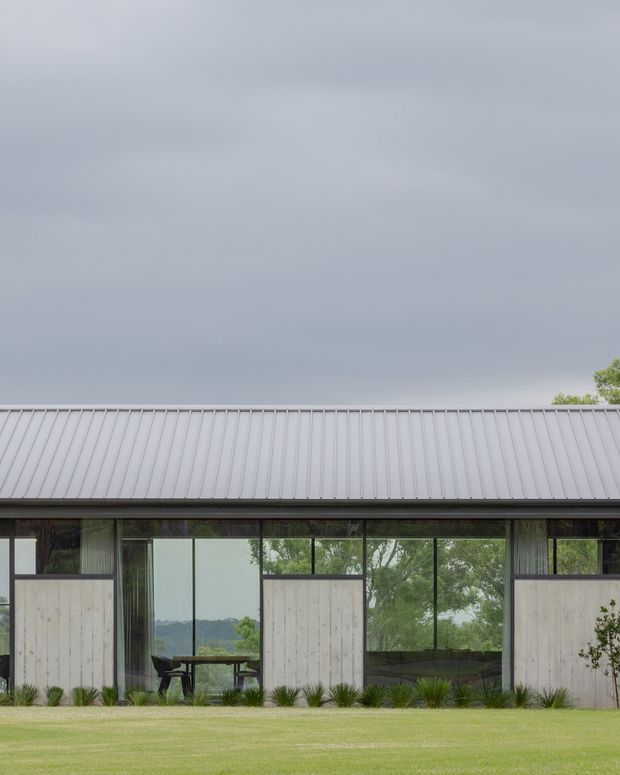 A low, modern single-storey building viewed from the landscaped lawn. The facade combines extensive dark-framed glazing with pale vertical-panel sections, beneath a long pitched roof clad in light grey standing-seam metal. Interior furnishings are visible through the windows, and ornamental grasses line the building edge. The setting appears green and spacious under an overcast sky.