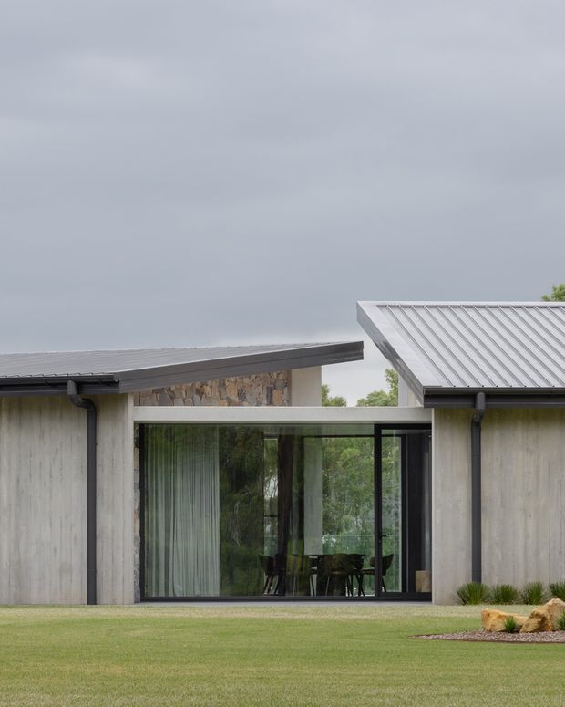 Exterior view of a contemporary single-storey residence composed of low-pitched roofed concrete volumes connected by a glazed central link. The facade features textured grey concrete walls, dark metal roof edging and gutters, expansive black-framed glazing, and a stone-clad section. A manicured lawn and restrained landscaping sit in the foreground beneath an overcast sky.