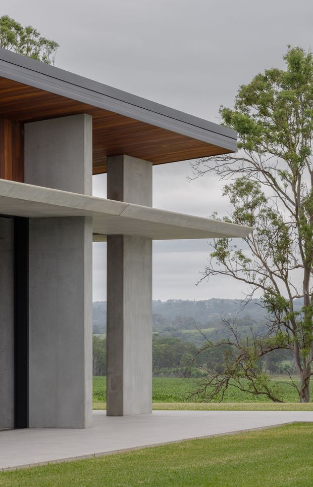 Exterior architectural photograph of a minimalist concrete house with strong horizontal cantilevered slabs, tall rectangular concrete columns, dark timber soffits, and a flat roof edge. The open facade frames a rural, tree-filled landscape and rolling hills under an overcast sky.