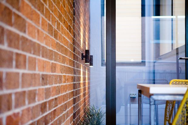 Close-up view of a contemporary dining area beside an exposed red brick wall, with black-framed glazing, a timber dining table, and bright yellow woven dining chairs. The image emphasizes the textural brickwork and indoor-outdoor visual connection.