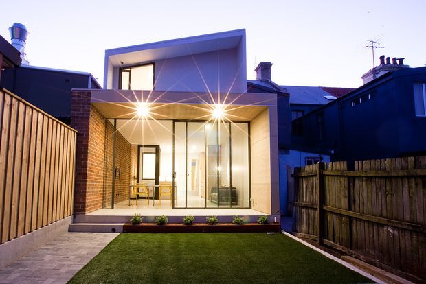Rear exterior view of a compact contemporary extension or pavilion opening onto a small landscaped courtyard. The design features a projecting upper volume, large floor-to-ceiling glazed doors, exposed red brick side walls, pale rendered framing, a paved path, and a rectangular lawn enclosed by timber fencing. Warm interior lighting contrasts with the cool evening exterior.