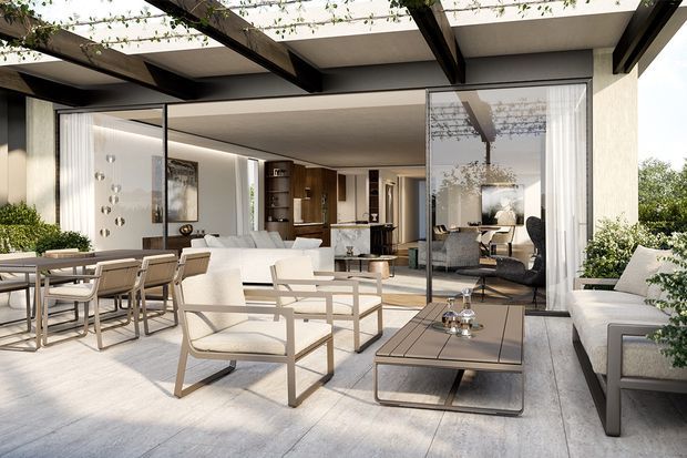 A bright, expansive covered outdoor terrace with pale stone or concrete-look flooring, a dark timber or metal pergola structure, and large glazed sliding doors opening into a refined contemporary living area. The terrace is furnished with light upholstered lounge chairs, a sofa, a dining table and chairs, and a low rectangular coffee table. Climbing greenery overhead and planting around the edges soften the clean architectural setting.