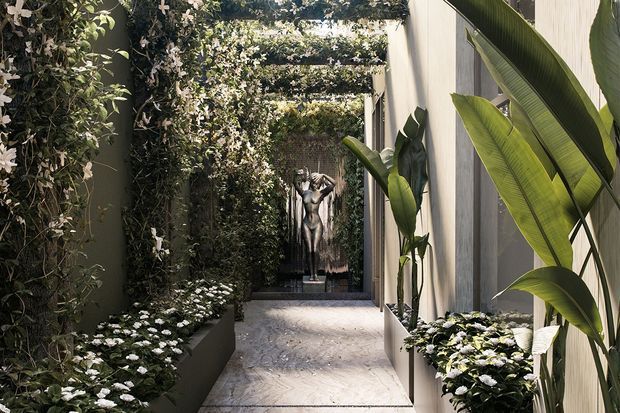 A narrow, refined landscaped courtyard walkway framed by dense vertical greenery and flowering plants. Pale textured walls and tall tropical foliage line the passage, while a series of overhead plant-covered trellises creates a shaded, tunnel-like effect. A classical human sculpture forms a focal point at the far end, with light stone or concrete paving underfoot.