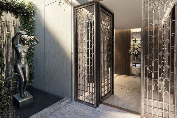 Elegant residential entrance with ornate, open wrought-iron security gates and a dark timber-framed doorway. A sculptural figure and vertical greenery occupy a small courtyard niche to the left, while the entry hall beyond features warm timber wall panelling, a light carpeted floor, floral decor, and softly illuminated interiors. The space has a refined, luxurious character with strong decorative metalwork.