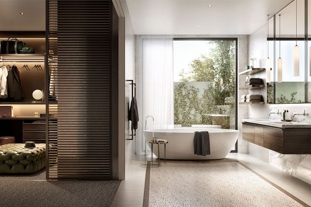 A refined contemporary bathroom with a freestanding white bathtub positioned beside large floor-to-ceiling glazing, a floating timber vanity with twin basins, and pendant lights. A walk-in wardrobe with illuminated shelving and hanging clothing is visible through a wide opening on the left. The room uses pale stone or tile surfaces, warm timber joinery, dark slatted screening, and a textured neutral rug, creating a luxurious, calm atmosphere.