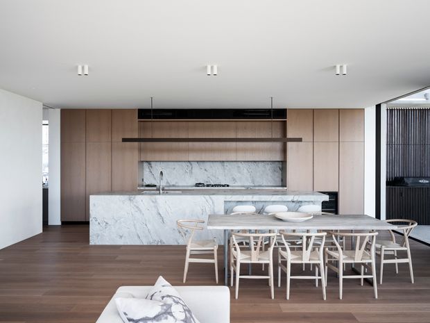 Open-plan contemporary kitchen and dining area with full-height light timber cabinetry, a large veined stone island and splashback, dark timber flooring, and a pale timber dining table with matching chairs. Minimal white ceiling with recessed downlights and dark-framed glazing at the right create a clean, refined interior.
