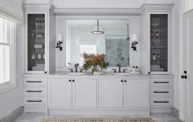 A bright, symmetrical bathroom vanity with a large white built-in cabinet, double sinks, dark hardware, and glass-front storage towers on either side. Decorative wall sconces, a central pendant light, pale wall panelling and trim, a large mirror, and a patterned rug create a refined traditional interior.