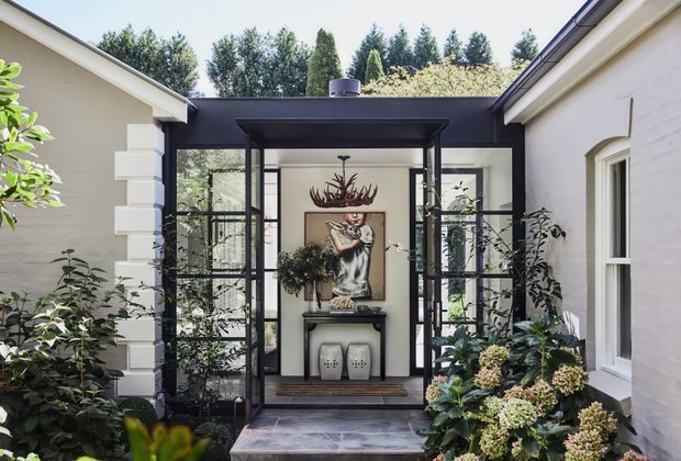 A refined courtyard entry framed by two pale rendered wings, with a black steel-and-glass pavilion forming a glazed entrance vestibule. The entry features dark tiled flooring, a console table, decorative artwork, plants, and layered garden planting, creating a formal yet inviting transition space.