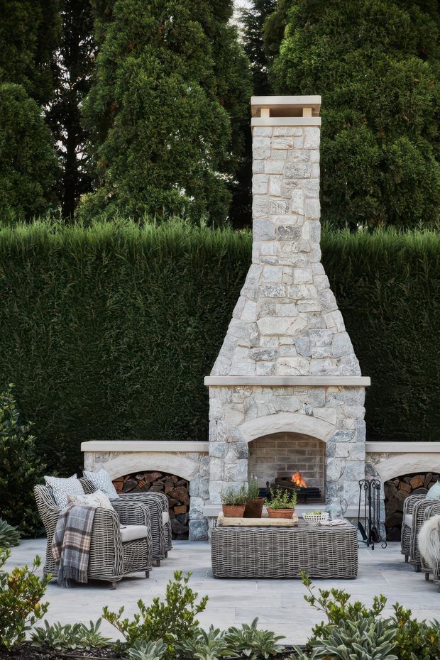 An elegant outdoor entertaining area centred on a tall, pale natural-stone fireplace chimney and arched firebox. The fireplace is integrated into low stone walls with built-in firewood storage, while wicker lounge chairs, a woven coffee table, cushions and throws create a comfortable seating arrangement on a pale paved terrace. Dense clipped hedges, topiary-like trees and layered planting form a lush garden backdrop.