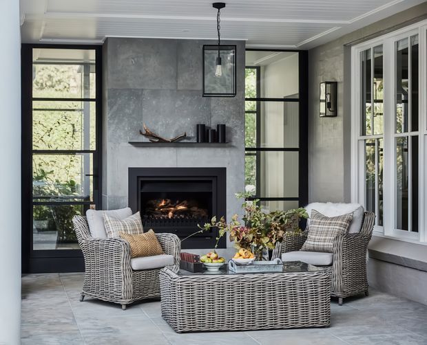 A sophisticated enclosed sitting room or sunroom with a central fireplace, grey concrete-look feature wall, large black-framed glazed doors and windows, woven wicker lounge furniture, and a pale panelled ceiling. The room has a calm, refined indoor-outdoor character with a neutral grey, white and natural tan palette.