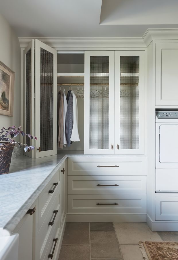 A bright, elegant laundry room with custom white built-in cabinetry, upper cupboards featuring fine wire-mesh glass panels, open hanging storage for clothing, and a stacked washer-dryer. The room includes a pale veined stone countertop, traditional recessed-panel drawer fronts, dark metal hardware, and large neutral stone-look floor tiles. A framed artwork and potted plant add decorative detail.