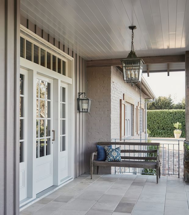 Covered residential front porch or veranda with a tiled floor, white French-style glazed entry doors, vertical striped wall cladding, a painted brick side wall, timber ceiling lining, dark hanging and wall-mounted lanterns, and a timber bench with cushions. The space opens toward a neatly clipped hedge and paved area beyond.
