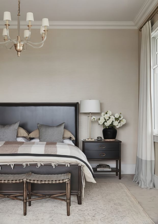 A refined bedroom interior with a dark upholstered and timber-framed bed, layered neutral bedding, a black bedside chest, table lamp, floral arrangement, woven bench, pale patterned rug, full-height curtains, and a large window. The room has beige textured wallcovering, white decorative ceiling moulding, and a restrained neutral palette with dark wood and black accents.