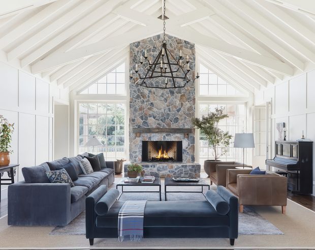 Bright, spacious living room with a vaulted white timber-beamed ceiling, white wall panelling, large multi-pane windows, and a prominent natural irregular-stone fireplace chimney breast. The room is furnished with deep blue sofas and ottoman seating, brown leather armchairs, a dark coffee table, a traditional metal chandelier, and a piano. The overall scheme is refined and comfortable, combining coastal Hamptons-inspired detailing with rustic stone and classic furnishings.