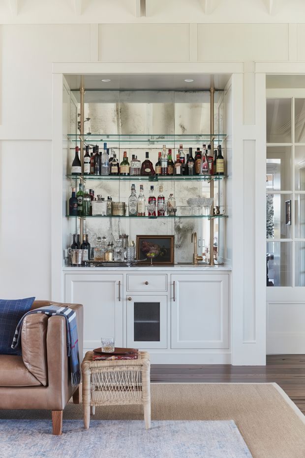 A bright, refined living room featuring a built-in home bar with white panelled cabinetry, mirrored backing, glass shelves, brass supports, and a stone or dark benchtop. The bar is stocked with bottles and glassware. A leather armchair with a blue patterned throw, woven stool, layered rugs, and dark timber flooring are visible in the foreground. Glazed white-framed doors open to an adjoining space.