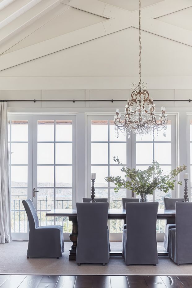 Bright, elegant dining room with a long dark wood dining table surrounded by upholstered slipcovered chairs. Large multi-pane French doors and windows line the wall, providing abundant natural light and views beyond. The room features a dramatic crystal chandelier, white-painted timber wall and ceiling panelling, decorative curtains, and a large leafy floral arrangement. A raised light carpeted dining platform transitions to dark timber flooring in the foreground.